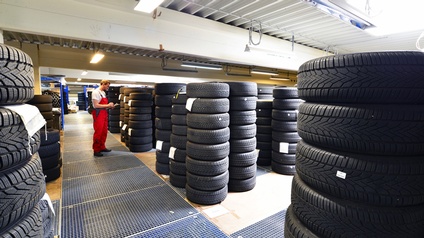 Person in roter Arbeitslatzhose in Autoreifenlager stehend nimmt Notizen auf Clipboard vor, ringsum aufgetürmte Autoreifen