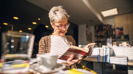 Lächelnde Person mit Brillen sitzt an Tisch und hält Buch in Händen auf das sie blickt, im Hintergrund verschwommen Tische mit Bücherstapeln und volle Bücherregale