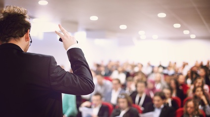 Rückenansicht einer Person, die auf einer Bühne steht und spricht. Der Person gegenüber sitzen viele verschiedene Personen auf Sesseln in mehreren Reihen