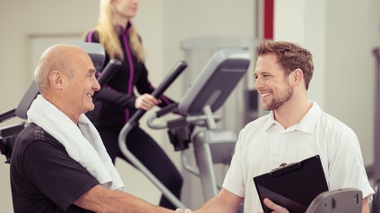 Person schüttelt einer älteren Person mit einem Handtuch auf den Schultern die Hand. Im Hintergrund ist eine weitere Person auf einem Steppgerät.