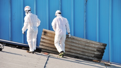 Zwei Personen mit weißen Schutzanzügen und Helmen sowie Atemschutzmasken in Rückenansicht auf Wellblechdach vor blauer Wand stehend, eine Person hievt Wellblechpanel
