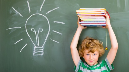 Kind hält Bücherstapel über Kopf, im Hintergrund Kreidetafel mit aufgemalter Glühbirne