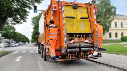 Zwei Personen in orange-reflektierenden Jacken und Hosen stehen hinten auf einem Müllauto und halten sich fest. Das Müllauto fährt auf einer Straße. Im Hintergrund sind parkende Autos, Gebäude und eine Allee.