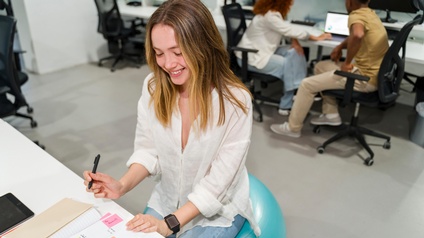 Eine jugendliche Person mit offenen, hellen Haaren sitzt auf einem Sportball an einem Schreibtisch in einem Raum mit mehreren Schreibtischen. Vor ihr ist ein aufgeklapptes Notizbuch, in dem verschiedene Post-its kleben. Die Person hält einen Stift