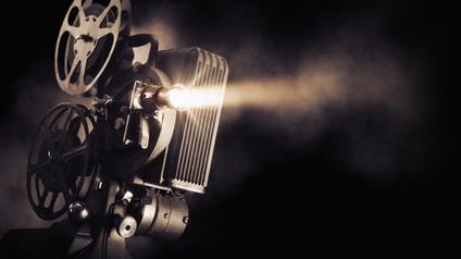 Ein Filmprojektor vor schwarzem Hintergrund, von dem ein heller Lichtschein ausgeht.