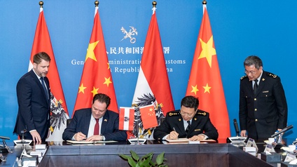 Zwei Personen in Anzügen sitzen hinter einem Tisch und halten jeweils einen Stift in der rechten Hand, den sie auf ein Blatt Papier halten. Hinter ihnen sind die Flaggen Österreichs und Chinas