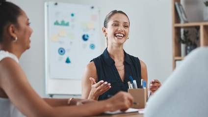 Zwei Personen sitzen nebeneinander an einem Tisch in einem Büro. Beide lachen. Auf dem Tisch sind Zettel sowie Stifte. Hinter der einen Person steht ein Whiteboard mit Zettel mit verschiedenen Grafiken und Statistiken