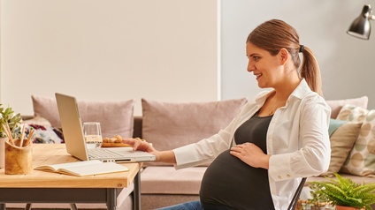 Schwangere Person sitzt freudig bei einem Wohnzimmertisch und arbeitet mit einem Laptop während im Hintergrund eine Couch sowie Pflanzen stehen