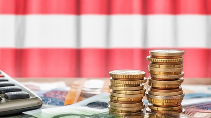 Im Vordergrund sind zwei Münzstapel, unter denen verschiedene Geldscheine liegen. In der linken Bildhälfte ist ein Taschenrechner. Dahinter hängt eine Flagge in den Farben rot-weiß-rot
