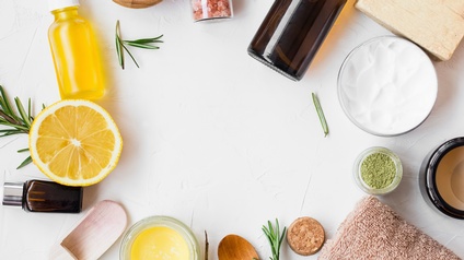 Verschiedene Objekte zum Thema Kosmetik wie Honig, braune Fläschchen, grünes Pulver, ein kleiner Holzlöffel, ein Handtuch, gelbe Flüssigkeit in einem durchsichtigem Fläschchen sind in einem Kreis auf weißem Hintergrund angeordnet.