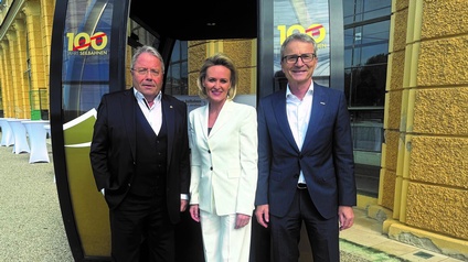 Porträt von drei Personen nebeneinander vor einer Gondel, die vor einem Gebäude steht. Alle tragen Businesskleidung. Auf beiden Seiten der Gondel ist ein Logo mit der Aufschrift „100 Jahre Seilbahnen“ angebracht