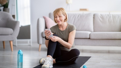 Person mittleren Alters mit schulterlangen blonden Haaren sitzt mit Fitnesskleidung auf einer Fitnessmatte und blickt dabei auf ein Smartphone während sich im Hintergrund ein heller Wohnraum mit Couch befindet