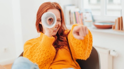 Person mit roten langen Haaren und gelbem Pullover blickt durch eine zusammengefaltete Rolle Papier und zeigt in die Kamera