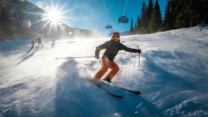 Eine Person mit Helm, Schutzbrille, Skianzug, Skistöcken und Skiern befindet sich parallel zur schneebedeckten Piste und hebt die Arme leicht an. Im Hintergrund sind weitere Personen mit Skiern. Über ihnen ist ein Sessellift.