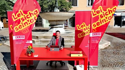 Mann sitzt an rotem Schreibtisch auf einem Platz in einer Stadt mit großen roten Bannern herum.