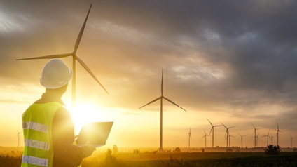 Person mit Warnweste und weißem Schutzhelm hält einen Laptop in Händen und blickt auf einen Windpark bei Sonnenuntergang