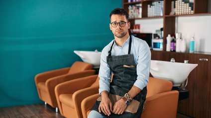 Person mit Schürze sitzt in einem modernen Friseursalon; im Hintergrund leere Waschplätze und Stühle.