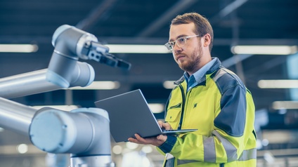 Person in Schutzkleidung und Brille verwendet einen Laptop bei der Arbeit an einem Roboticarm, neue Ära in einer automatisierten Produktionsstätte