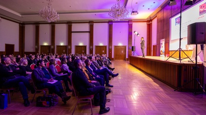 Publikum in einem Veranstaltungsraum verfolgt eine Präsentation auf einer Bühne mit Leinwand.