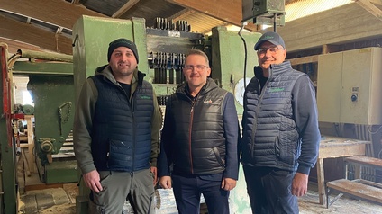 Drei Personen stehen in einer Werkhalle vor einer großen grünen Industriemaschine auf einem mit Sägespänen bedeckten Holzboden.