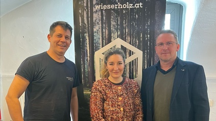 Drei Personen stehen in einem Innenraum vor einem Roll-up-Banner mit Waldmotiv und der Aufschrift „wieserholz.at“.