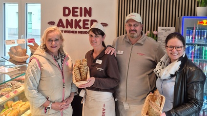 Vier Personen stehen in einer Bäckereifiliale vor einer Verkaufstheke und halten Körbe mit länglichen, bestreuten Gebäckstücken in den Händen; im Hintergrund sind Backwaren, ein Getränkekühlschrank und das Logo „DEIN ANKER“ zu sehen, Gesichter sind unkenntlich gemacht.