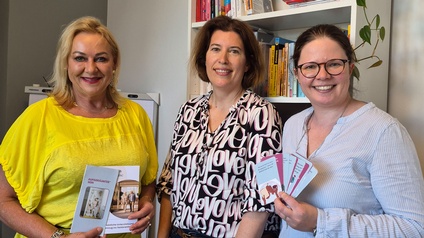 Drei Personen stehen in einem Büro vor einem Regal mit Büchern und Pflanze und halten Broschüren in den Händen.