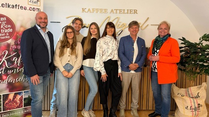 Gruppenfoto von mehreren Personen in einem Kaffee-Atelier vor einer Wand mit der Aufschrift „Kaffee Atelier“, daneben ein Roll-up mit Kaffeebohnen-Motiv und Markenhinweis.