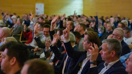 Eine große Gruppe von Menschen sitzt in einem Veranstaltungssaal und hebt gleichzeitig die Hand um abzustimmen. Die Teilnehmenden tragen überwiegend Business-Kleidung und befinden sich in einem gut beleuchteten Raum mit Holzvertäfelung.