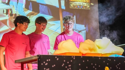 Drei Personen in pinken T‑Shirts führen auf einer Bühne bei der Science Night 2026 ein naturwissenschaftliches Experiment durch. Vor ihnen steigen große gelbe Schaummengen aus mehreren Behältern auf. Im Hintergrund sind Projektionen und Bühnenelemente zu sehen.
