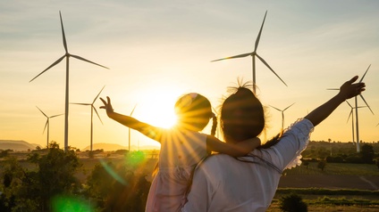 Person mit Kind am Arm schaut in den Sonnenuntergang - am Horizont stehen Windräder.