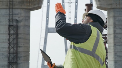 Person in Seitenansicht in gelber Warnweste, mit Helm und Schutzbrillen blickt nach oben und deutet mit Hand in Blickrichtung, in anderer Hand Tablet haltend, im Hintergrund verschwommen Betonpfeiler einer Brücke