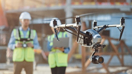 Fliegende Drohne im Fokus, im Hintergrund stehen zwei Personen mit weißen Schutzhelmen und gelber Warnweste in der Unschärfe und übernehmen die Steuerung