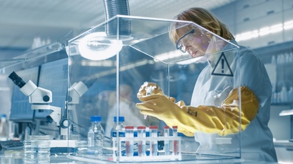 Eine Person mit Schutzkleidung arbeitet in einem hochmodernen Hightech-Labor mit Computern und Analysegeräten und untersucht mit Chemikalien befüllte Reagenzgläser, die sich in einer Handschuhbox befinden.
