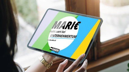 Fokus auf Tablet, das Person vor Fenster in Händen hält, auf Display in verschiedenen Farben umrahmtes Sujet mit schwarzem Schriftzug auf weißem kreisrund ausgespartem Hintergrund Marie mehr.wert bei Unternehmertum marie.wko.at