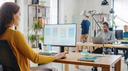 Im Fokus Person in Rückenansicht vor Computerbildschirm in Großraumbüro sitzend und Computermaus haltend, im Hintergrund weitere Personen sitzend und stehend