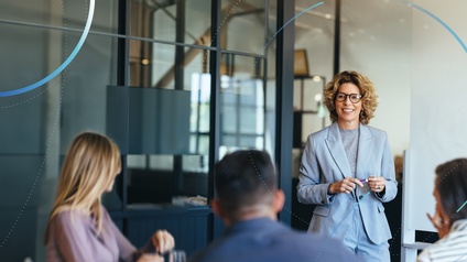 Eine Person im hellen Anzug mit Brille steht lächelnd vor einer kleinen Gruppe  in einem modernen Besprechungsraum und hält einen Stift in der Hand, als würde sie etwas erklären.