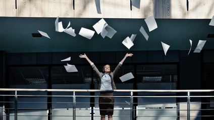Eine Person im Business-Outfit steht auf einer Galerie in einem modernen Gebäude, wirft zahlreiche Papierblätter jubelnd in die Luft und streckt dabei beide Arme nach oben.