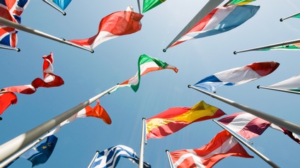 Mehrere Nationalflaggen aus verschiedenen Ländern wehen an hohen Masten im Wind vor einem klaren blauen Himmel