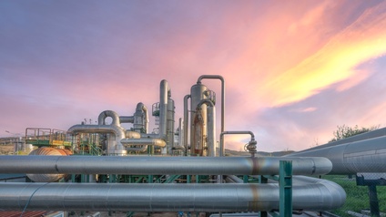 Vor rötlichem Himmel verlaufen im Vordergrund Pipelines. Dahinter sind weitere Pipelines sowie Silos und Metalltreppen. Rechts ist eine grüne Wiese