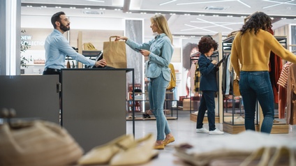 In einem Raum sind mehrere Kleiderstangen, an den jeweils andere Kleidungsstücke auf Haken hängen. Vor manchen stehen Personen. Eine Person steht einer Person an einem erhöhten Tisch gegenüber. Zwischen den beiden steht eine braune Einkaufstasche