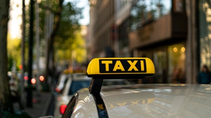 Nahaufnahme des hellen Dachs eines Autos. Auf dem Dach ist ein gelb-leuchtendes Objekt montiert, auf dem Taxi steht. Vor diesem Auto stehen weitere Autos. Rechts neben dem Auto ist ein Gebäude
