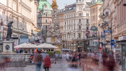 Der Graben in Wien mit seinen historischen Gebäuden sowie zwei Statuen wie der Pestsäule. In der Fußgängerzone gehen und stehen Menschen