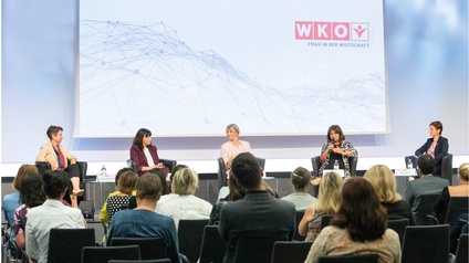 Mehrere Person auf Podium sitzend vor Leinwand mit WKO Logo und Schriftzug Frau in der Wirtschaft, im Vordergrund Publikum in Rückenansicht