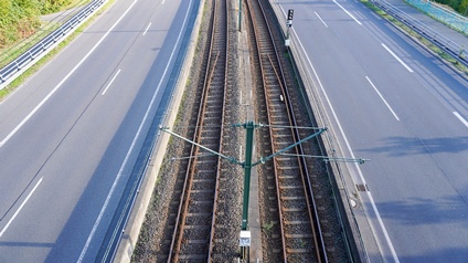 In der Mitte sind zwei Gleise parallel nebeneinander. Links und rechts von den Gleisen ist jeweils eine mehrspurige Straße