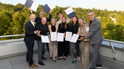 9 Personen stehen auf Terrasse und halten freudig Urkunden hoch