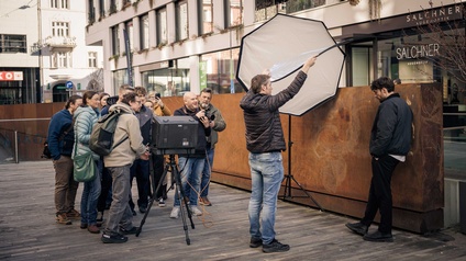 Szene aus einem Fotoshooting, mehrere Personen sind zu sehen. Man sieht links einen Fotografen und rechts lehnt ein Mann an einer Wand, der in dieser Position fotografiert werden soll