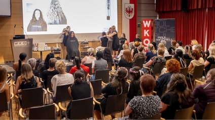 Veranstaltungssaal mit einer Bühne. Viele Menschen sitzen im Publikum. Vorne ist eine Bühne, auf dieser sitzen zwei Personen, die gerade die Haare geschnitten bekommen.