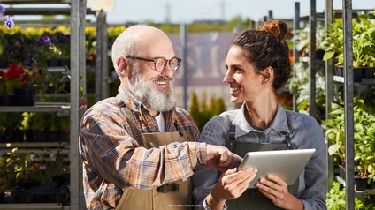 Nahaufnahme von zwei Personen, die in einer Gärtnerei nebeneinander stehen und sich ansehen. Die rechte Person hat einen Bart und eine Brille und deutet auf das Tablet, das die linke Person in den Händen hält. Im Hintergrund Metallregale mit Pflanzen