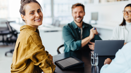 Personen sitzen am Tisch im Büro, haben ein Tablet vor sich liegen und eine Person blickt direkt in die Kamera
